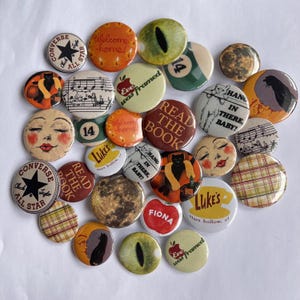 May include: A collection of round buttons with various designs and text. Some buttons feature a Converse All Star logo, a black cat, musical notes, and the text "READ THE BOOK". Other buttons have faces, an eye, and the text "Welcome home!"