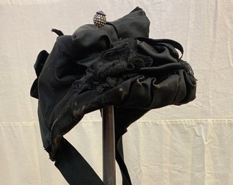 Puede incluir: Un sombrero de terciopelo negro con ribete de encaje negro y una cinta negra. El sombrero está en un soporte y tiene un pequeño adorno plateado en la parte superior.