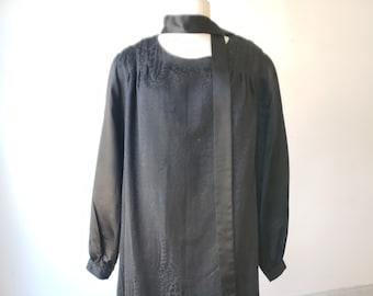 Op de afbeelding: Een zwarte jurk met een lange mouw, loszittende top en een aansluitende rok. De top heeft een patroon en een zwarte strik om de nek.