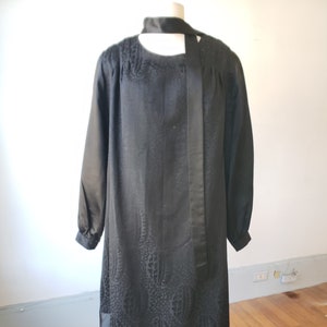 May include: A black dress with a long-sleeved, loose-fitting top and a fitted skirt. The top has a patterned design and a black ribbon tie at the neck.