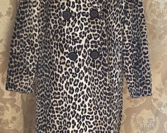 Puede incluir: Un abrigo de doble botonadura con estampado de leopardo, cuello ancho y cuatro botones negros. El abrigo se exhibe en un maniquí y tiene mangas largas. El patrón del abrigo presenta manchas negras sobre un fondo beige.