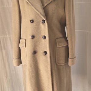 May include: A long, tan, double-breasted coat featuring a wide notched collar, six dark brown buttons arranged in two vertical rows, and two front flap pockets. The coat has long sleeves with folded cuffs and a textured, wool-like fabric finish.