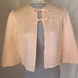 May include: A pale peach quilted jacket featuring a diamond-stitched pattern, three-quarter length sleeves, and a round neckline. The front opening is secured with two matching ribbon ties positioned at the upper chest area.