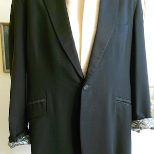 Puede incluir: Un blazer negro con solapa de satén y cierre de un solo botón. Las mangas están dobladas para revelar un forro estampado. Una bufanda color crema está drapeada en el interior. El blazer se exhibe en un maniquí.