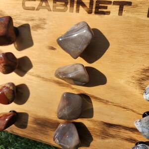 Pu&ograve; includere: Un pannello di legno con la parola "CABINET" bruciata sulla superficie. Il pannello ha sei pomelli di pietra attaccati ad esso. I pomelli sono di varie tonalit&agrave; di marrone e grigio.