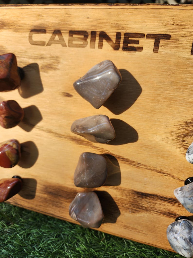 Pu&ograve; includere: Un pannello di legno con la parola "CABINET" bruciata sulla superficie. Il pannello ha sei pomelli di pietra attaccati ad esso. I pomelli sono di varie tonalit&agrave; di marrone e grigio.