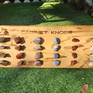 Może przedstawiać: Drewniana deska z napisem "CABINET KNOBS" umieszczonym na niej. Deska jest wypełniona różnymi gałkami z naturalnego kamienia w różnych kolorach i wzorach.
