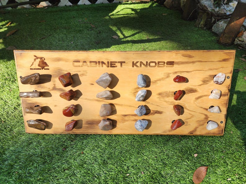 Pu&ograve; includere: Un pannello di legno con il testo "CABINET KNOBS" visualizzato su di esso. Il pannello &egrave; pieno di diversi pomelli in pietra naturale di diversi colori e motivi.