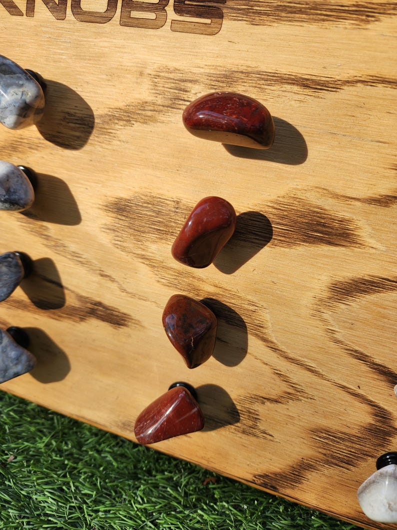 Pu&ograve; includere: Un pannello di legno con una finitura bruciata, che mostra una variet&agrave; di pomelli in pietra rossa e grigia. Il pannello ha la parola "Knobs" incisa su di esso.