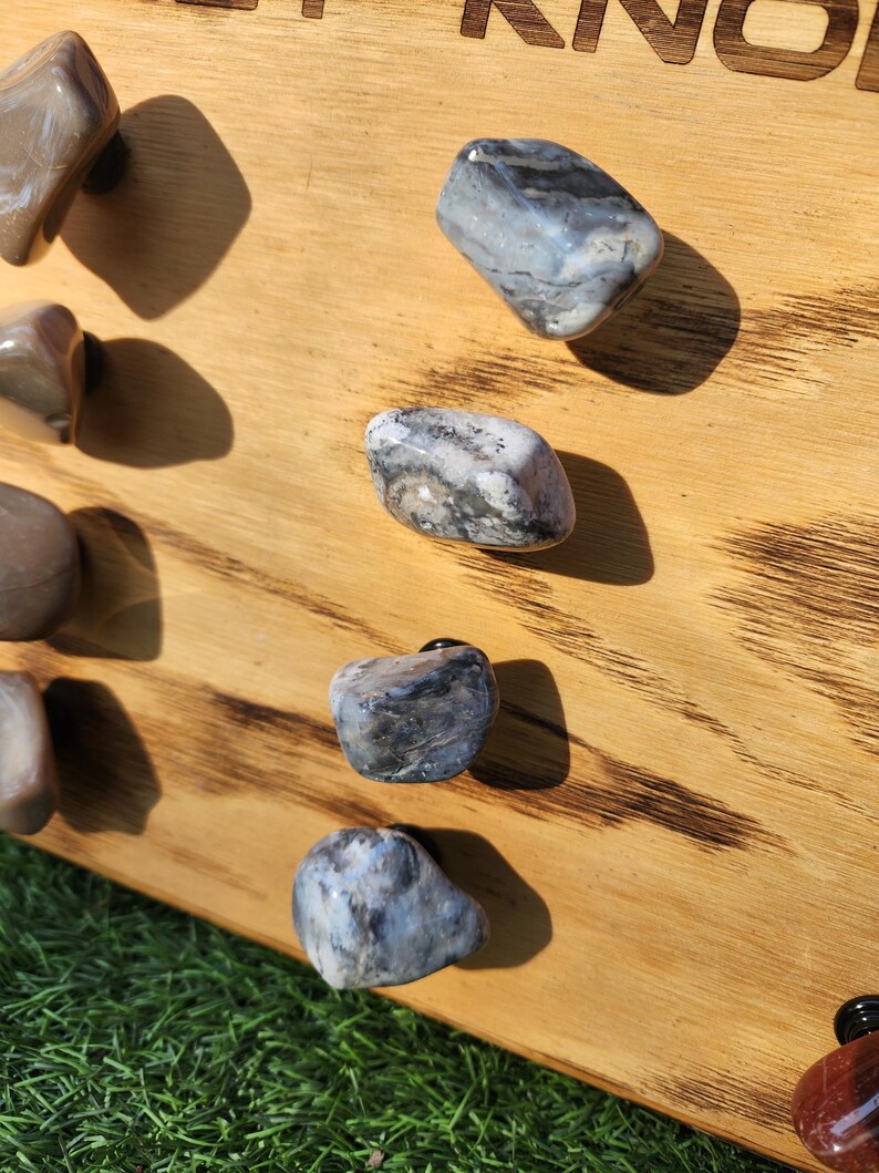 Pu&ograve; includere: Primo piano di sei pomelli in pietra naturale, ognuno con un motivo e un colore unici, montati su una tavola di legno. I pomelli sono di colore grigio chiaro con striature nere e bianche.