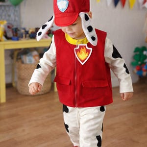 Op de afbeelding: Een kinderkostuum met een rode brandweer-stijl vest en hoed met een pootafdruk-embleem. Het kostuum bevat een witte broek en een shirt met lange mouwen met zwarte vlekken en rode schoenen. De outfit is ontworpen voor een themafeest.