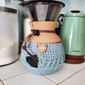 Puede incluir: Una funda de ganchillo azul claro para cafetera con un botón de madera y detalles de cordón de cuero. La cafetera es de vidrio y corcho. Una tetera verde está en el fondo.
