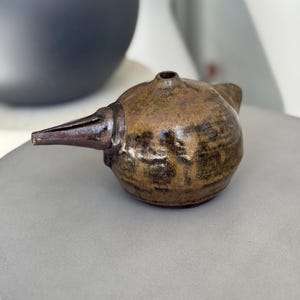 Puede incluir: Un pequeño jarrón de cerámica marrón con un pico en forma de pájaro. El jarrón tiene un esmalte texturizado y moteado con acentos marrones y negros.