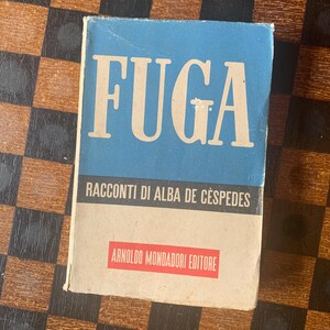 May include: A vintage book titled "FUGA" with a light blue cover and white text. The book also features the text "RACCONTI DI ALBA DE CESPEDES" and "ARNOLDO MONDADORI EDITORE" in a red rectangle. The book is on a checkered surface.