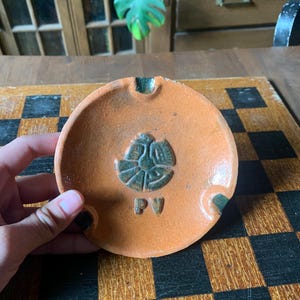 以下が含まれることがあります： 丸いオレンジ色の陶器の灰皿。濃い緑色のエンブレムと「PV」の文字がエンボス加工されています。灰皿には、タバコを置くための濃い緑色の溝が2つあります。手作りの灰皿で、素朴な外観をしています。