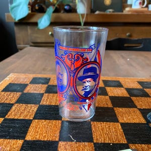 May include: A clear glass featuring a red, white, and blue design with portraits of Winston Churchill. The glass has the text "Winston Churchill" and "Wings Across the Sea". The glass is on a checkered surface.