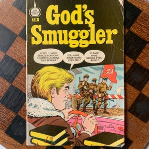 Puede incluir: Cómic vintage titulado "God's Smuggler" con una portada amarilla y negra. La portada presenta a un hombre rubio conduciendo un coche rosa y soldados con una bandera roja. El texto incluye "Spire Christian Comics" y "39¢".