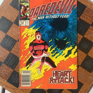 以下が含まれることがあります： デアデビルをフィーチャーしたヴィンテージコミック。表紙には「DAREDEVIL THE MAN WITHOUT FEAR！」と「HEART ATTACK！」の文字。表紙のアートは、デアデビルが赤と黒のスーツを着て、燃えるようなオレンジと青の背景に描かれています。