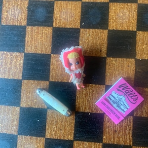May include: A small doll with a red hood and a white lace collar, a small pocket knife, and a pink matchbook with the text "Walt's" and "4680 EAGLEROCK BLVD." are arranged on a checkered surface.