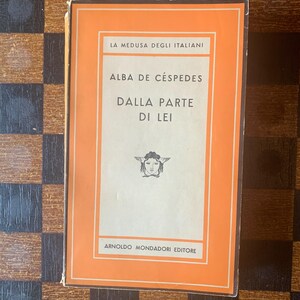 May include: A vintage book titled "Dalla Parte Di Lei" by Alba de Céspedes. The book has an orange cover with white text and a graphic of a woman's face. The publisher is Arnoldo Mondadori Editore.