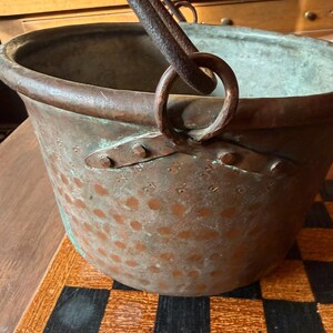May include: Antique copper cauldron with a riveted handle and a large, circular opening. The cauldron has a weathered patina with green and brown tones, and is set on a checkered surface. The handle is made of iron.