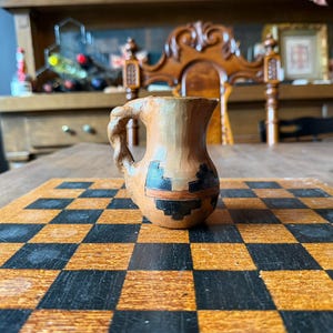 May include: A small, light brown ceramic pitcher featuring a braided handle and a decorative band of black geometric patterns encircling the lower body. The vessel has a rounded base and a flared rim, showcasing a smooth, matte-like finish.