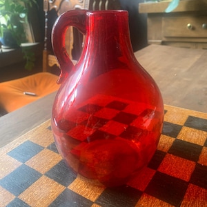 Puede incluir: Una jarra de vidrio rojo brillante con asa. La jarra tiene un cuerpo redondeado y un patrón de cuadros. La jarra está sobre una superficie de madera.