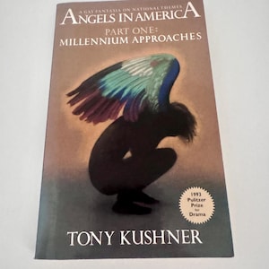 Op de afbeelding: Een paperbackboek getiteld Angels in America Part One: Millennium Approaches door Tony Kushner. De omslag toont een donkere, gehurkte figuur met grote, levendige blauwe en groene vleugels tegen een bruine achtergrond en een zegel van de Pulitzerprijs 1993.