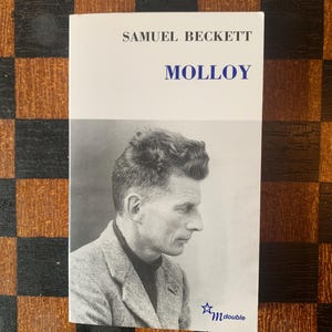 Puede incluir: Un libro de bolsillo titulado "Molloy" de Samuel Beckett. La portada presenta un retrato en blanco y negro de un hombre. El libro está sobre un fondo a cuadros.