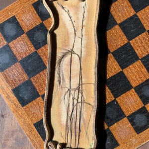 Puede incluir: Una bandeja rectangular de cerámica beige con un borde texturizado y tejido. La bandeja presenta un diseño botánico estilizado de hierbas altas y espigas de trigo en tonos marrones y negros. La bandeja está colocada sobre un tablero de ajedrez de madera.