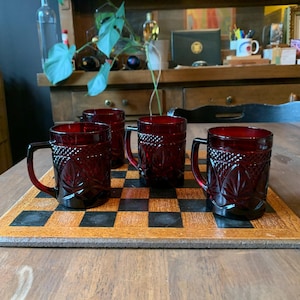 May include: Four ruby red glass mugs with a textured pattern and handles are arranged on a black and brown checkerboard. The mugs are on a wooden table, with a shelf and decor in the background.