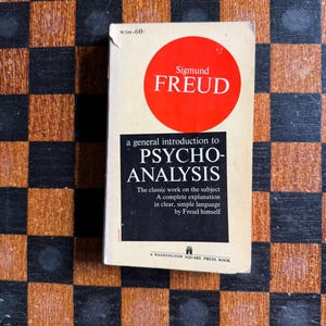 May include: A paperback book titled "A General Introduction to Psycho-Analysis" by Sigmund Freud. The book has a white cover with a red circle containing the author's name. The book is on a brown and black checkered surface.