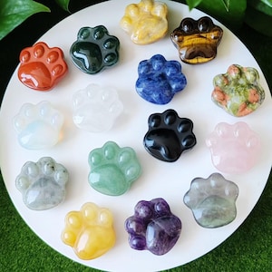 May include: A collection of 15 polished gemstone paw print charms in various colors, including red, green, blue, black, white, pink, and yellow. The charms are arranged on a white surface.