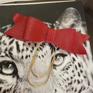 May include: A red leather bow-shaped paper clip with a gold-colored metal clip. The bow is textured and sits atop a black and white image of a leopard's face.