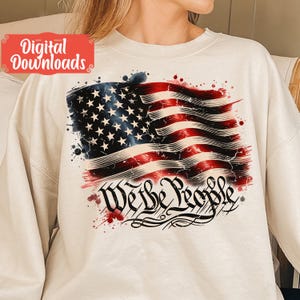 May include: A white sweatshirt featuring a distressed American flag design and the words "We the People" in a stylised font. The flag is red, white and blue with a watercolour effect.