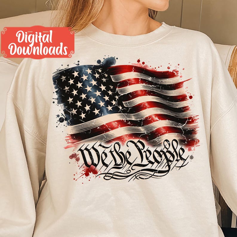 May include: A white sweatshirt featuring a distressed American flag design and the words "We the People" in a stylised font. The flag is red, white and blue with a watercolour effect.