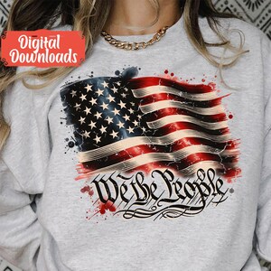 May include: A grey sweatshirt featuring a distressed American flag design and the words "We the People" in a stylised font. The flag is red, white and blue with white stars on a blue field. There are red, white and blue paint splatters around the flag.