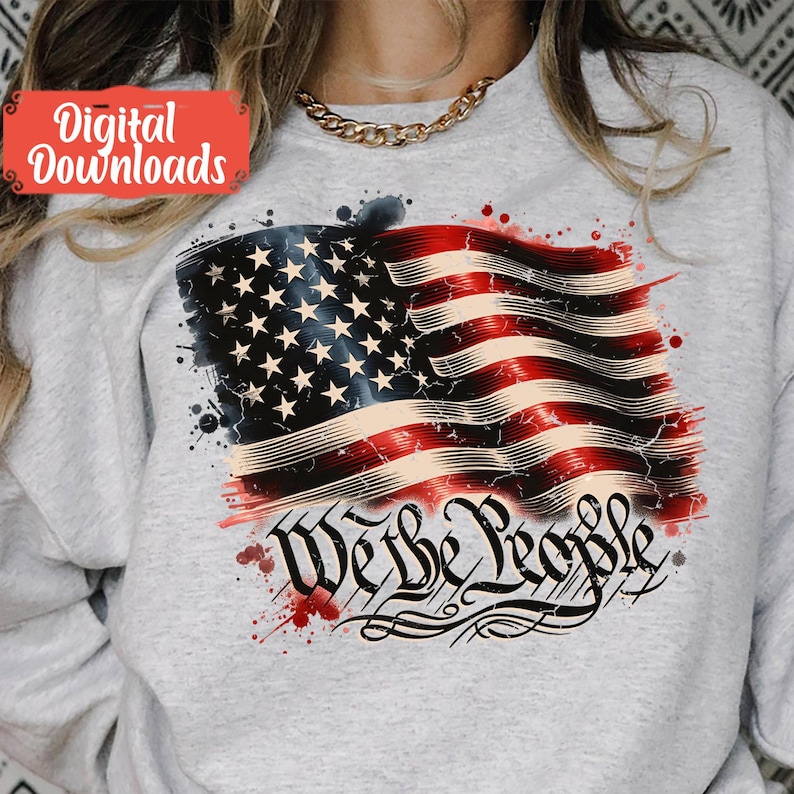 May include: A grey sweatshirt featuring a distressed American flag design and the words "We the People" in a stylised font. The flag is red, white and blue with white stars on a blue field. There are red, white and blue paint splatters around the flag.