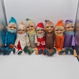 May include: A collection of seven seated gnome dolls with smiling faces, each wearing a different colored outfit and hat. The dolls have white beards and are holding small accessories like a shovel and a broom. The dolls are approximately 6 inches tall.