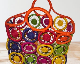 May include: A colorful woven tote bag with a circular design. The bag features a vibrant array of colors, including red, orange, yellow, green, and blue. The handles are a bright orange, and the bag is made of a woven material.