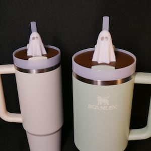 May include: Two stainless steel travel mugs with lids and handles. The mug on the left is light pink and the mug on the right is light green. Both mugs have a ghost-shaped lid with a straw.
