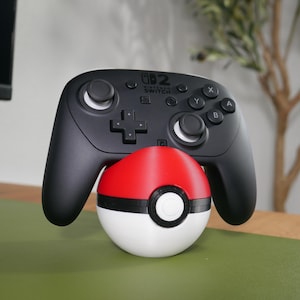 May include: A black Nintendo Switch Pro controller resting on a spherical red and white stand designed to resemble a Poké Ball. The controller features standard analog sticks, a directional pad, and buttons labeled A, B, X, and Y on the front face.