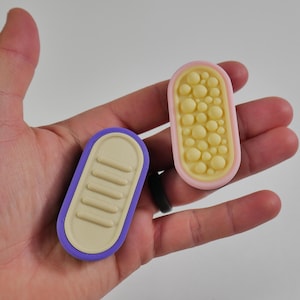 Op de afbeelding: Twee kleine, ovale objecten worden in een hand gehouden. Eén heeft een paarse rand met een beige interieur en horizontale ribbels. De andere heeft een roze rand met een gele binnenkant gevuld met kleine, ronde bobbels. De objecten lijken decoratief of functioneel.