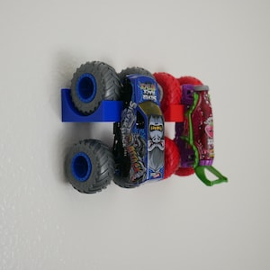 Pode incluir: Um suporte de parede de plástico azul segurando um brinquedo de caminhão monstro azul e prateado com um gráfico de gorila e o texto Silver Back Attack, ao lado de um suporte vermelho com um caminhão monstro roxo e vermelho com detalhes verdes.