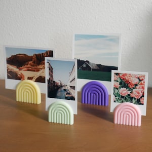 以下が含まれることがあります： カラフルな虹型のホルダーに飾られた、ポラロイド風の写真のコレクション。ホルダーは黄色、緑、紫、ピンクの色合いです。写真は風景や花の配置を描いており、装飾的なディスプレイを作成しています。