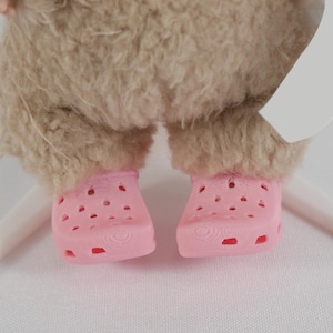 May include: Close-up of a doll's feet wearing pink clogs. The clogs have multiple holes and a textured sole. The doll's legs are covered in a light brown, fluffy material.