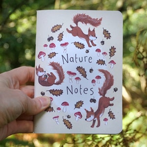 Könnte beinhalten: Ein cremefarbenes Notizbuch mit den Worten "Nature Notes" und Illustrationen von Eichhörnchen, Pilzen, Eicheln und Blättern. Das Notizbuch wird in einer Hand vor einem verschwommenen grünen Hintergrund gehalten.