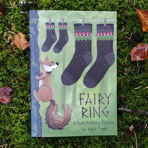 May include: A book cover titled "Fairy Ring" featuring a whimsical illustration of socks hanging on a clothesline. A cartoon fox clings to a tree trunk. The socks are dark gray with mushroom and green accents. The background is green.