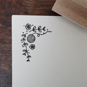 May include: A black ink floral corner design featuring a ball of yarn, printed on white paper. A wooden stamp is in the upper right corner. The design includes flowers, leaves, and dots, creating a decorative border.