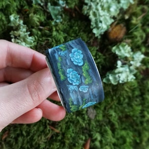 May include: Washi tape with a dark blue background and a watercolour design of light blue flowers and green foliage. The tape is held in a hand against a backdrop of green moss.